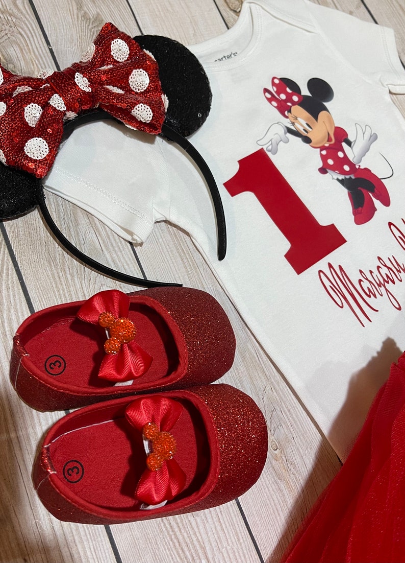 May include: A red and white Minnie Mouse themed outfit for a baby girl. The outfit includes a white onesie with a red number one and Minnie Mouse graphic, a red tulle skirt, a red glitter headband with a red and white polka dot bow, and red glitter shoes with a Minnie Mouse bow.