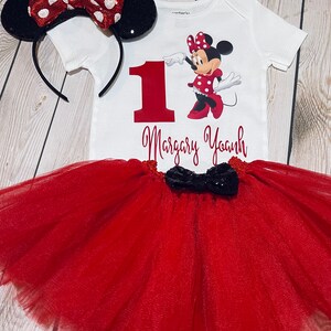 May include: A red and white Minnie Mouse themed first birthday outfit with a white bodysuit, red tulle skirt, and black bow. The bodysuit features a red number one, a graphic of Minnie Mouse, and the name "Margary Yoanh".
