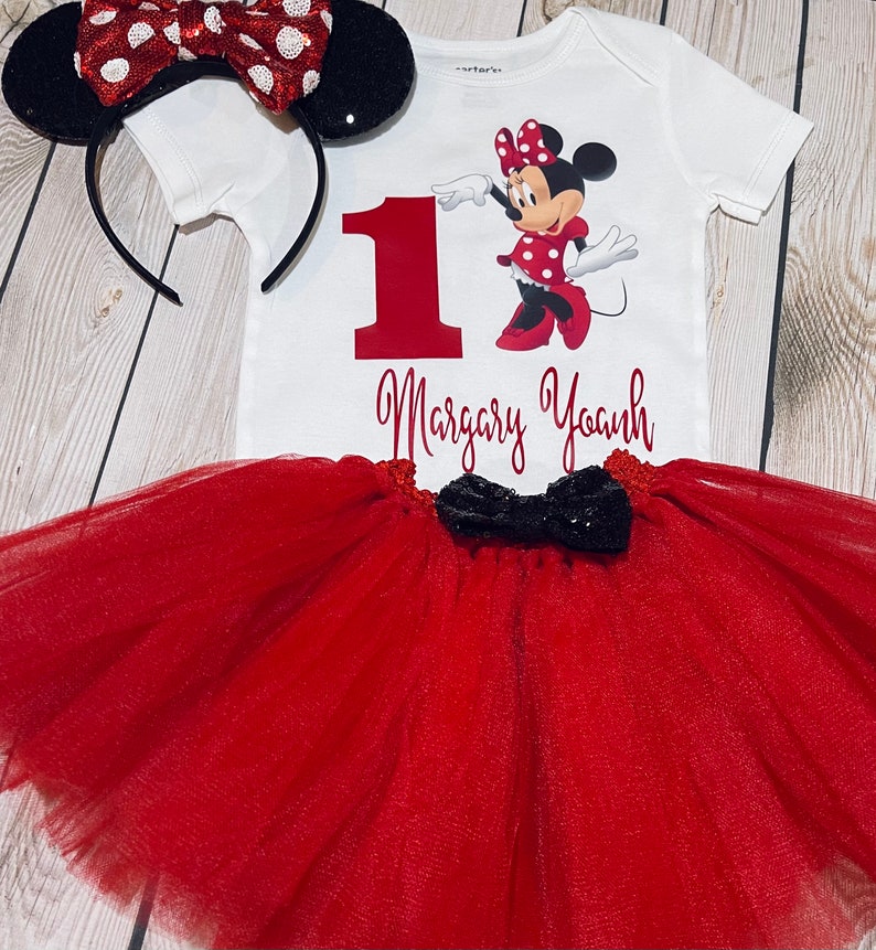 May include: A red and white Minnie Mouse themed first birthday outfit with a white bodysuit, red tulle skirt, and black bow. The bodysuit features a red number one, a graphic of Minnie Mouse, and the name "Margary Yoanh".