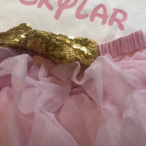 Puede incluir: Un tut&uacute; de tul rosa con un lazo de lentejuelas doradas. El tut&uacute; se combina con un body blanco con el n&uacute;mero "1" y el nombre "SKYLAR" en rosa.