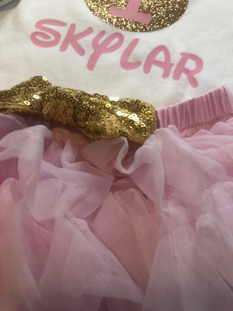 Puede incluir: Un tut&uacute; de tul rosa con un lazo de lentejuelas doradas. El tut&uacute; se combina con un body blanco con el n&uacute;mero "1" y el nombre "SKYLAR" en rosa.