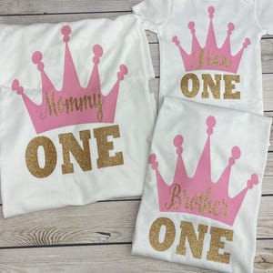 May include: Three white t-shirts with pink crowns and gold glitter text. The shirts say "Mommy One", "Brother One", and "One".