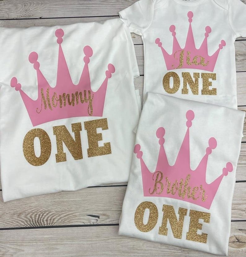 May include: Three white t-shirts with pink crowns and gold glitter text. The shirts say "Mommy One", "Brother One", and "One".