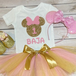 May include: A pink and gold tutu with a gold bow, a white onesie with a gold glitter Minnie Mouse head and the number 1, and a pink glitter headband with Minnie Mouse ears. The onesie also says "BAJA" in pink.
