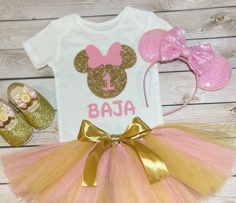 May include: A pink and gold tutu with a gold bow, a white onesie with a gold glitter Minnie Mouse head and the number 1, and a pink glitter headband with Minnie Mouse ears. The onesie also says "BAJA" in pink.