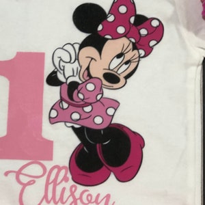 May include: A white onesie with a pink number one and the name "Ellison" printed on it. A pink and white Minnie Mouse character is printed on the onesie.