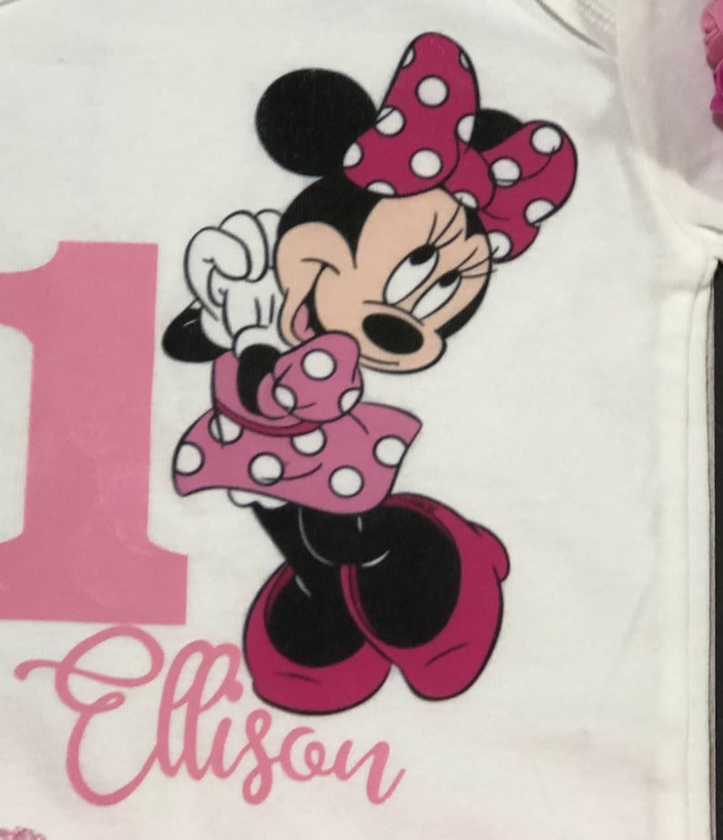 May include: A white onesie with a pink number one and the name "Ellison" printed on it. A pink and white Minnie Mouse character is printed on the onesie.