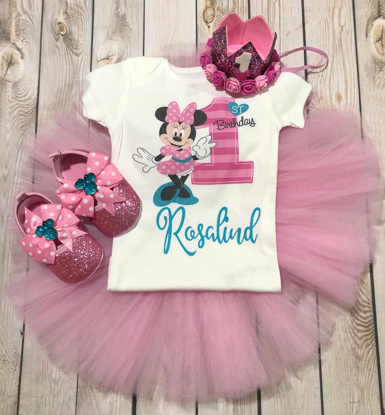 Może przedstawiać: R&oacute;żowa sp&oacute;dniczka tiulowa, biały body z numerem jeden w r&oacute;żowo-białe paski, grafiką Minnie Mouse oraz tekstem "1. urodziny" i "Rosalind". Body ma r&oacute;wnież r&oacute;żową kokardkę na kołnierzyku. Str&oacute;j uzupełnia r&oacute;żowa korona z srebrną jedynką i r&oacute;żowymi kwiatami.