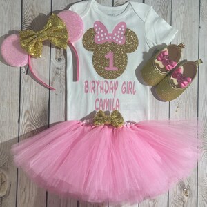 May include: Pink and gold Minnie Mouse themed first birthday outfit for a baby girl. The outfit includes a white onesie with a gold glitter Minnie Mouse head, the number one, and the text "Birthday Girl Camila", a pink tulle tutu with a gold bow, a pink headband with gold glitter ears, and gold glitter shoes with pink bows.