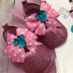 Może przedstawiać: R&oacute;żowe błyszczące buciki dla niemowląt z białymi kokardkami w kropki i turkusową ozdobą w kształcie głowy Minnie Mouse.
