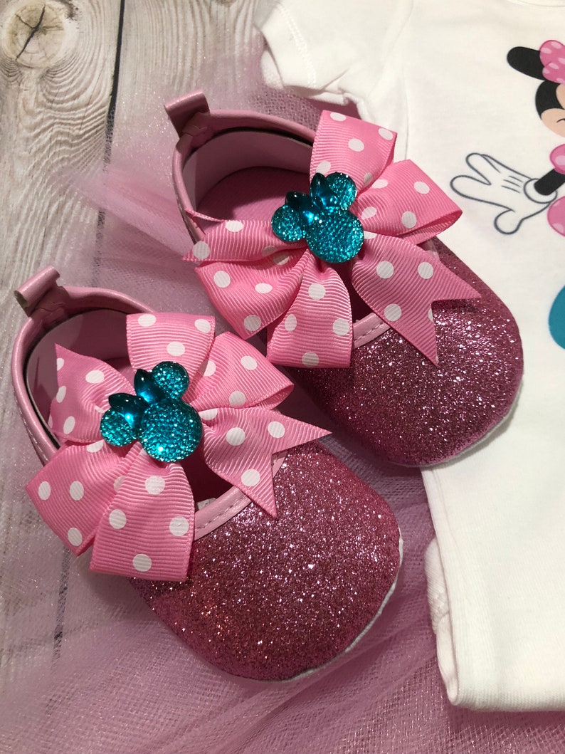 Może przedstawiać: R&oacute;żowe błyszczące buciki dla niemowląt z białymi kokardkami w kropki i turkusową ozdobą w kształcie głowy Minnie Mouse.