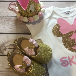 Puede incluir: Un body blanco con una cabeza de Minnie Mouse de purpurina dorada y un lazo rosa, el texto "Skylar" en purpurina rosa y el n&uacute;mero "1" en purpurina dorada. Una corona de purpurina dorada con fieltro rosa y flores blancas y rosas est&aacute; encima del body. Un par de zapatos de purpurina dorada con lazos rosas y orejas de Minnie Mouse est&aacute;n en el body. El body tiene una etiqueta que dice "carter's 18m".