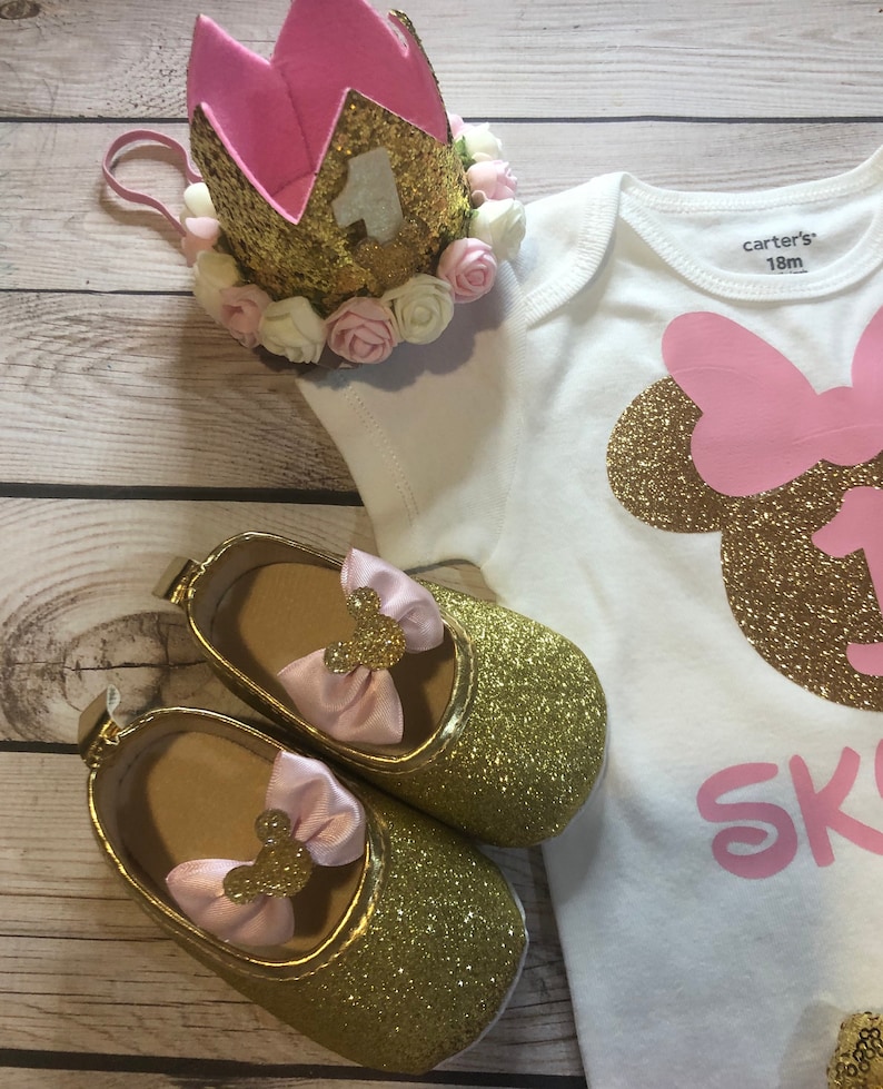 Puede incluir: Un body blanco con una cabeza de Minnie Mouse de purpurina dorada y un lazo rosa, el texto "Skylar" en purpurina rosa y el n&uacute;mero "1" en purpurina dorada. Una corona de purpurina dorada con fieltro rosa y flores blancas y rosas est&aacute; encima del body. Un par de zapatos de purpurina dorada con lazos rosas y orejas de Minnie Mouse est&aacute;n en el body. El body tiene una etiqueta que dice "carter's 18m".