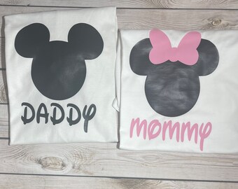 May include: Two white t-shirts with a black and white Mickey Mouse silhouette. The shirt on the left says "Daddy" in black and the shirt on the right says "Mommy" in pink.