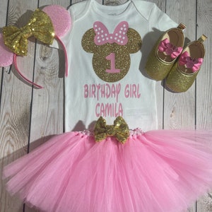 May include: A pink and gold Minnie Mouse themed first birthday outfit for a baby girl. The outfit includes a white bodysuit with a gold glitter Minnie Mouse head and the number one, a pink tulle tutu, a gold glitter headband with pink ears, and a pair of gold glitter shoes with pink bows.