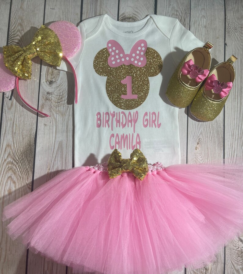 May include: A pink and gold Minnie Mouse themed first birthday outfit for a baby girl. The outfit includes a white bodysuit with a gold glitter Minnie Mouse head and the number one, a pink tulle tutu, a gold glitter headband with pink ears, and a pair of gold glitter shoes with pink bows.