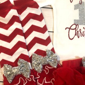 May include: A festive baby outfit featuring a white onesie with the text "Teagan 1st Christmas" in glittery red and silver. Includes red and white chevron leg warmers with silver sequin bows and a red tutu.