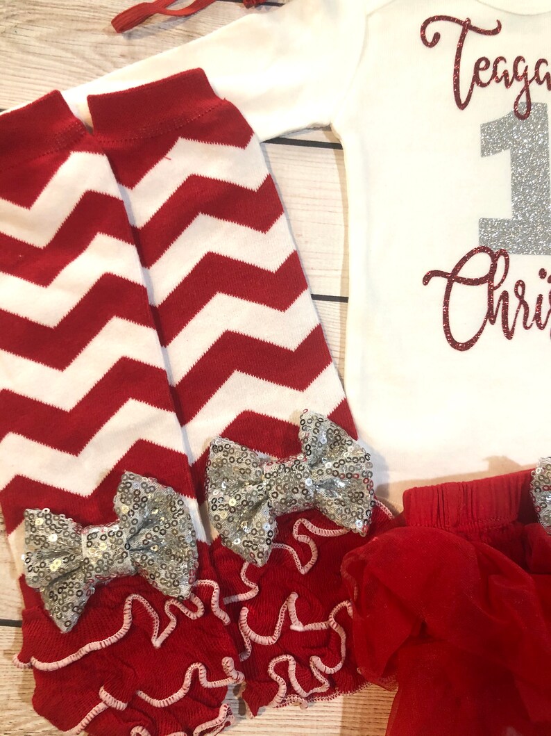 May include: A festive baby outfit featuring a white onesie with the text "Teagan 1st Christmas" in glittery red and silver. Includes red and white chevron leg warmers with silver sequin bows and a red tutu.