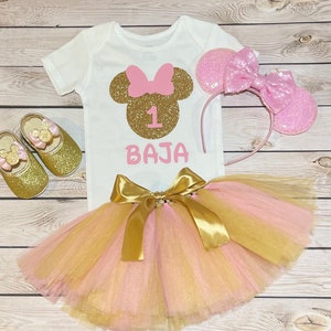 May include: A pink and gold tutu with a gold bow, a white onesie with a gold glitter Minnie Mouse head and the number 1, and a pink headband with ears. The onesie also has the name "BAJA" written on it.