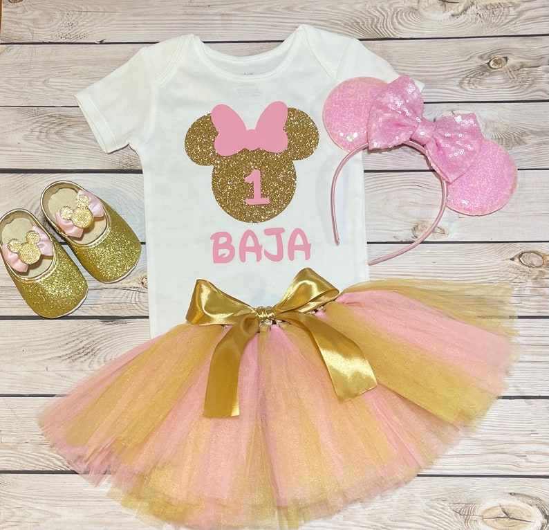 May include: A pink and gold tutu with a gold bow, a white onesie with a gold glitter Minnie Mouse head and the number 1, and a pink headband with ears. The onesie also has the name "BAJA" written on it.