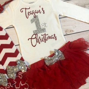 May include: A white long-sleeved baby bodysuit with the text "Teagan's 1st Christmas" in red glitter and silver glitter. The outfit includes a red tutu skirt, red and white chevron leggings, and a red headband.