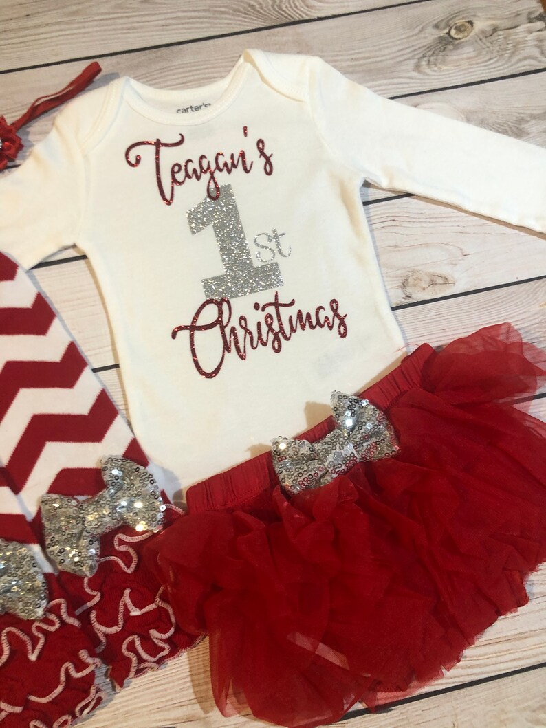 May include: A white long-sleeved baby bodysuit with the text "Teagan's 1st Christmas" in red glitter and silver glitter. The outfit includes a red tutu skirt, red and white chevron leggings, and a red headband.