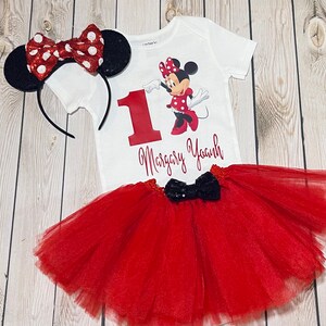 May include: A red and black Minnie Mouse themed first birthday outfit for a baby girl. The outfit includes a white bodysuit with a red number one, a red tulle tutu skirt, and a red and black Minnie Mouse headband.