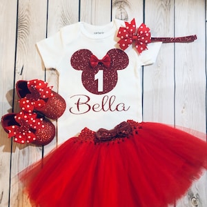 Puede incluir: Conjunto de primer cumplea&ntilde;os de Minnie Mouse rojo y blanco para una ni&ntilde;a. El conjunto incluye una camiseta blanca con una cabeza de Minnie Mouse roja de purpurina, el n&uacute;mero 1 y el nombre Bella. El conjunto tambi&eacute;n incluye una falda de tul roja y zapatos rojos de purpurina con lunares blancos.