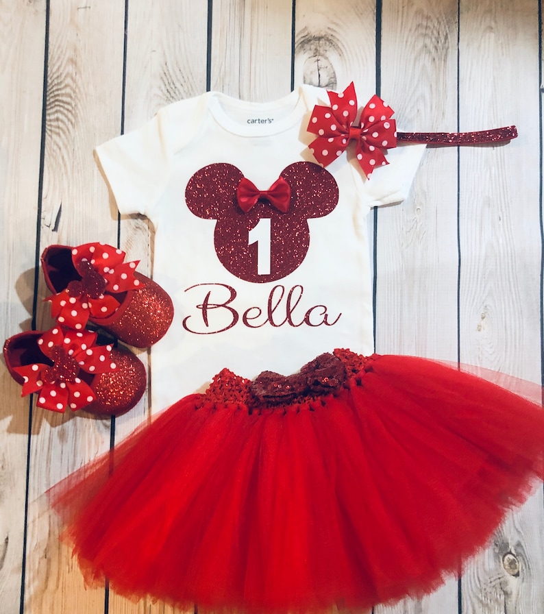 Puede incluir: Conjunto de primer cumplea&ntilde;os de Minnie Mouse rojo y blanco para una ni&ntilde;a. El conjunto incluye una camiseta blanca con una cabeza de Minnie Mouse roja de purpurina, el n&uacute;mero 1 y el nombre Bella. El conjunto tambi&eacute;n incluye una falda de tul roja y zapatos rojos de purpurina con lunares blancos.