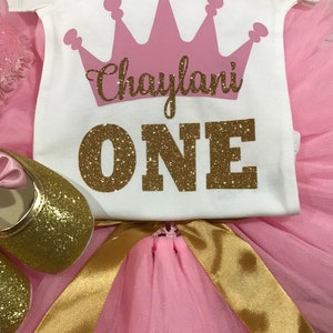 May include: Pink and gold tutu set for a first birthday. The white onesie has a pink crown and the text "Chaylani ONE" in gold glitter.