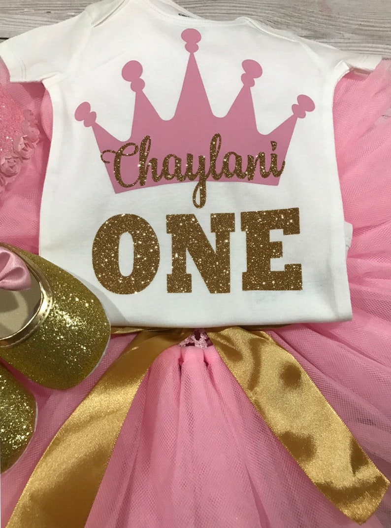 May include: Pink and gold tutu set for a first birthday. The white onesie has a pink crown and the text "Chaylani ONE" in gold glitter.