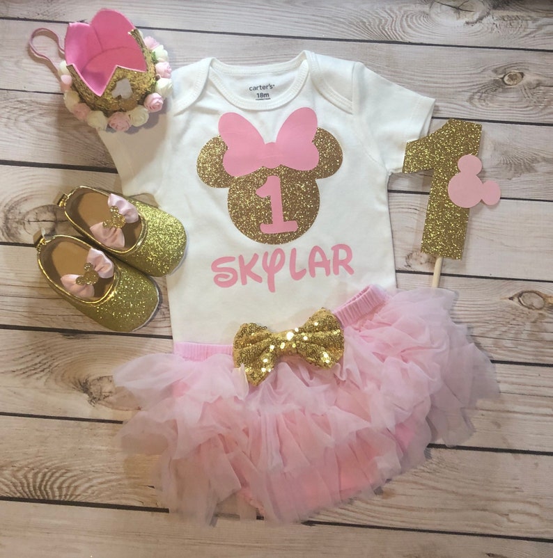 Puede incluir: Un conjunto de primer cumplea&ntilde;os rosa y dorado para una ni&ntilde;a. El conjunto incluye un body blanco con un n&uacute;mero uno de purpurina dorada y el nombre "SKYLAR" en purpurina rosa, una falda de tul rosa con un lazo de purpurina dorada y zapatos de purpurina dorada con lazos rosas. El conjunto tambi&eacute;n incluye una corona de purpurina rosa y dorada y un n&uacute;mero uno de purpurina dorada con una cabeza de Mickey Mouse rosa.
