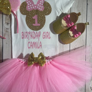 May include: A pink and gold Minnie Mouse themed first birthday outfit for a baby girl. The outfit includes a white bodysuit with a gold glitter Minnie Mouse head, a pink tutu, a gold bow, and gold shoes with pink bows.