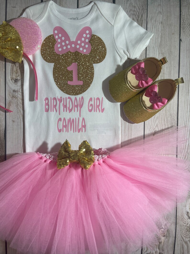 May include: A pink and gold Minnie Mouse themed first birthday outfit for a baby girl. The outfit includes a white bodysuit with a gold glitter Minnie Mouse head, a pink tutu, a gold bow, and gold shoes with pink bows.