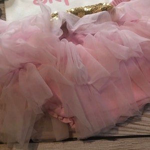 Puede incluir: Una falda de tut&uacute; de tul rosa con una cintura de lentejuelas rosa y dorada. La falda est&aacute; en capas y tiene un borde con volantes. La falda es para un beb&eacute; o un ni&ntilde;o peque&ntilde;o.