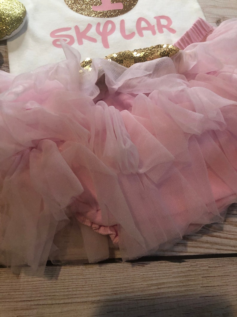 Puede incluir: Una falda de tut&uacute; de tul rosa con una cintura de lentejuelas rosa y dorada. La falda est&aacute; en capas y tiene un borde con volantes. La falda es para un beb&eacute; o un ni&ntilde;o peque&ntilde;o.