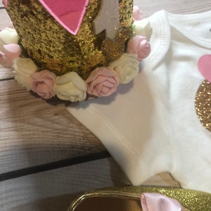 Puede incluir: Corona de purpurina dorada con forro de fieltro rosa y flores de tela blanca y rosa. La corona tiene un n&uacute;mero "1" de fieltro blanco.