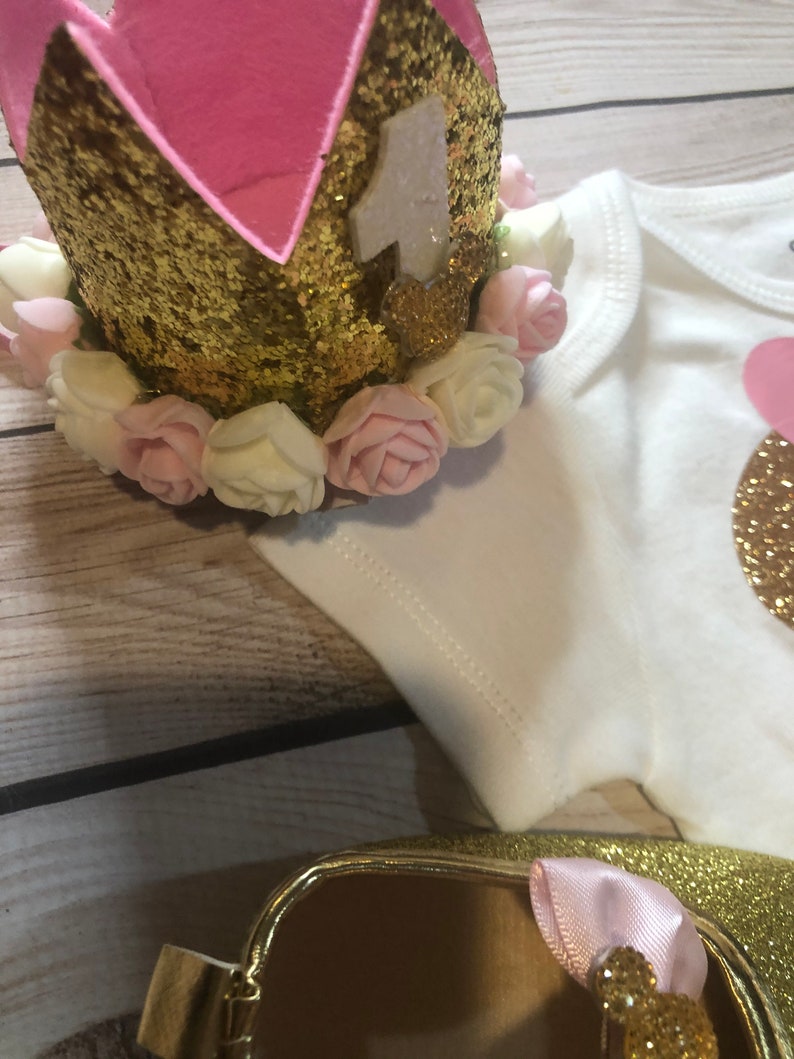 Puede incluir: Corona de purpurina dorada con forro de fieltro rosa y flores de tela blanca y rosa. La corona tiene un n&uacute;mero "1" de fieltro blanco.