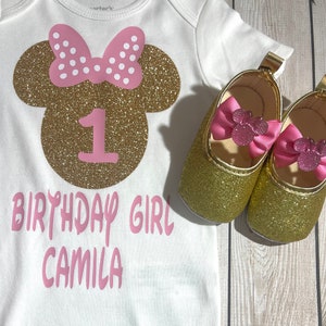 May include: A white onesie with a gold glitter Minnie Mouse head with pink polka dot bow and the number 1 in pink. The text "Birthday Girl Camila" is written in pink below the Minnie Mouse head. Two gold glitter shoes with pink bows are on a wooden surface.