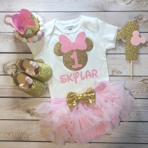 Puede incluir: Un conjunto de primer cumplea&ntilde;os de Minnie Mouse en rosa y dorado para una ni&ntilde;a. El conjunto incluye un body blanco con una cabeza de Minnie Mouse de purpurina dorada con un lazo rosa, el n&uacute;mero uno y el nombre "SKYLAR" en purpurina rosa. El conjunto tambi&eacute;n incluye un tut&uacute; de tul rosa con un lazo de purpurina dorada y zapatos de purpurina dorada.