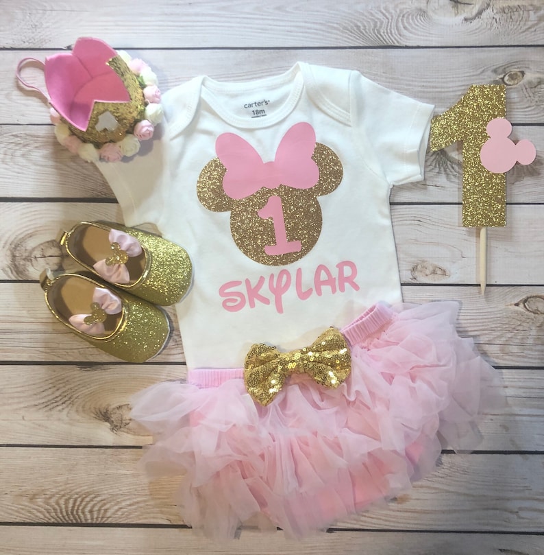 Puede incluir: Un conjunto de primer cumplea&ntilde;os de Minnie Mouse en rosa y dorado para una ni&ntilde;a. El conjunto incluye un body blanco con una cabeza de Minnie Mouse de purpurina dorada con un lazo rosa, el n&uacute;mero uno y el nombre "SKYLAR" en purpurina rosa. El conjunto tambi&eacute;n incluye un tut&uacute; de tul rosa con un lazo de purpurina dorada y zapatos de purpurina dorada.