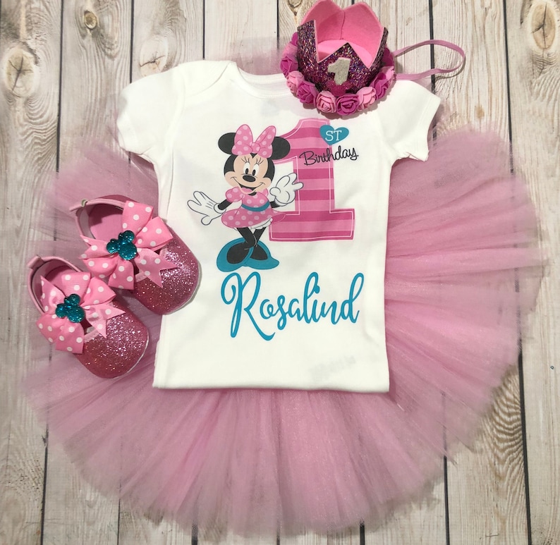 Może przedstawiać: R&oacute;żowa sp&oacute;dniczka tiulowa z białym body z grafiką Minnie Mouse, cyfrą 1 i tekstem "1. urodziny" i "Rosalind". Str&oacute;j uzupełnia r&oacute;żowa korona i r&oacute;żowe buty w białe kropki i turkusowe kokardki.
