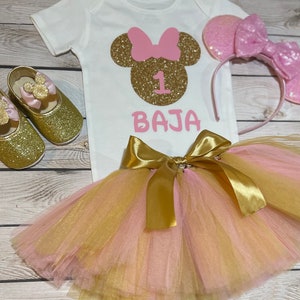 May include: A pink and gold Minnie Mouse themed first birthday outfit for a baby girl. The outfit includes a white onesie with a gold glitter Minnie Mouse head and the number 1, a pink and gold tulle tutu, a pink headband with glitter Minnie Mouse ears, and gold glitter shoes with pink bows.