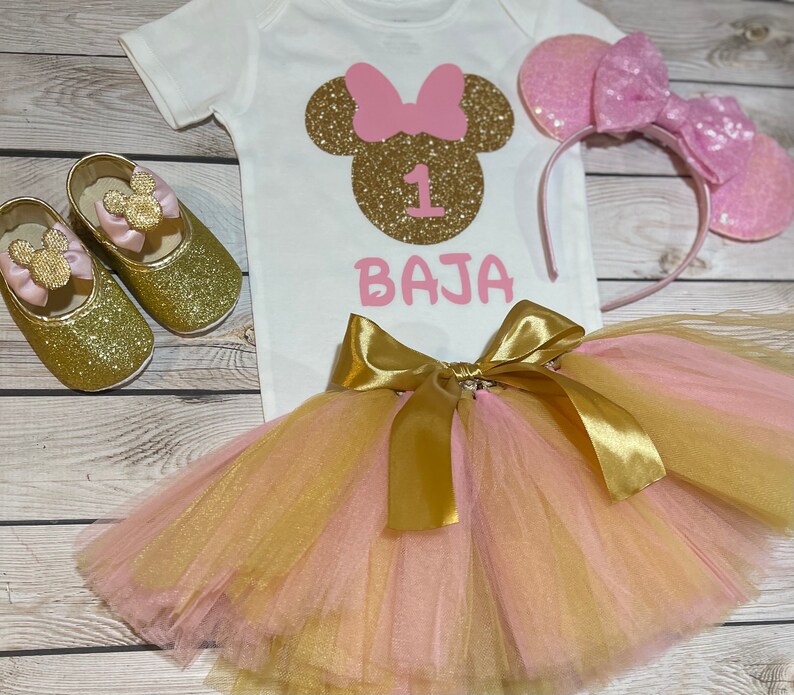 May include: A pink and gold Minnie Mouse themed first birthday outfit for a baby girl. The outfit includes a white onesie with a gold glitter Minnie Mouse head and the number 1, a pink and gold tulle tutu, a pink headband with glitter Minnie Mouse ears, and gold glitter shoes with pink bows.