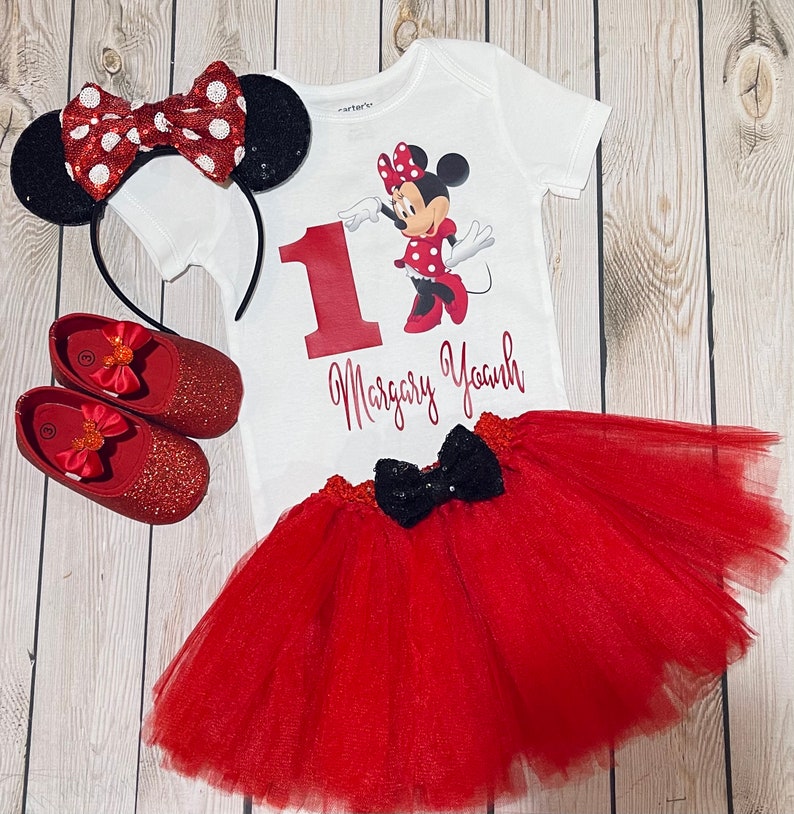 May include: A red and white Minnie Mouse themed first birthday outfit for a baby girl. The outfit includes a white bodysuit with a red number one, a red tulle tutu skirt, a red headband with black ears and a red bow, and red glitter shoes.