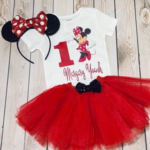 May include: A red and black Minnie Mouse themed first birthday outfit for a baby girl. The outfit includes a white bodysuit with a red number one, a red tulle tutu skirt, and a red and black Minnie Mouse headband.