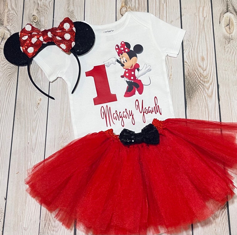 May include: A red and black Minnie Mouse themed first birthday outfit for a baby girl. The outfit includes a white bodysuit with a red number one, a red tulle tutu skirt, and a red and black Minnie Mouse headband.