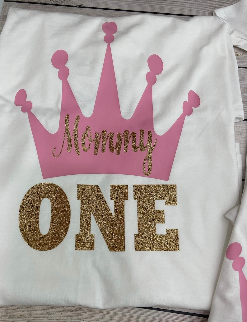 May include: White t-shirt with a pink crown and the words "Mommy One" in gold glitter.