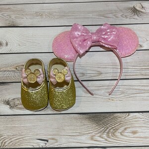 May include: Gold glitter baby shoes with pink bows and gold Mickey Mouse ears. The shoes have a pink bow on the front and the ears have a pink bow on top.