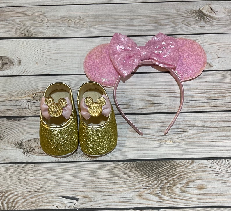May include: Gold glitter baby shoes with pink bows and gold Mickey Mouse ears. The shoes have a pink bow on the front and the ears have a pink bow on top.